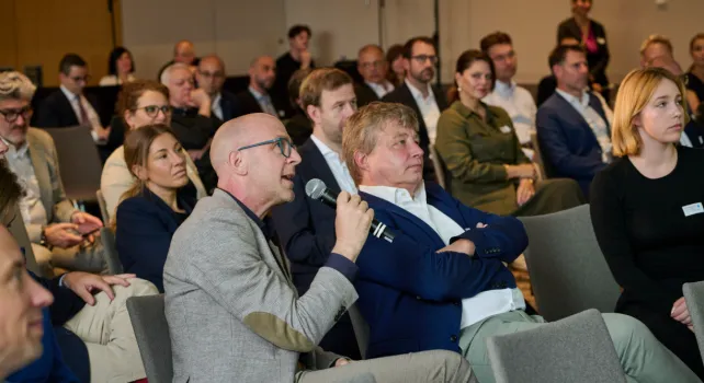 Spotlight Finance Basel Ein Saal voll sitzender Menschen, im Vordergrund ein Herr im grauen Anzug der sich mit einem Mikrofon zu Wort meldet.