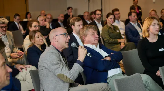 Ein Saal voll sitzender Menschen, im Vordergrund ein Herr im grauen Anzug der sich mit einem Mikrofon zu Wort meldet.