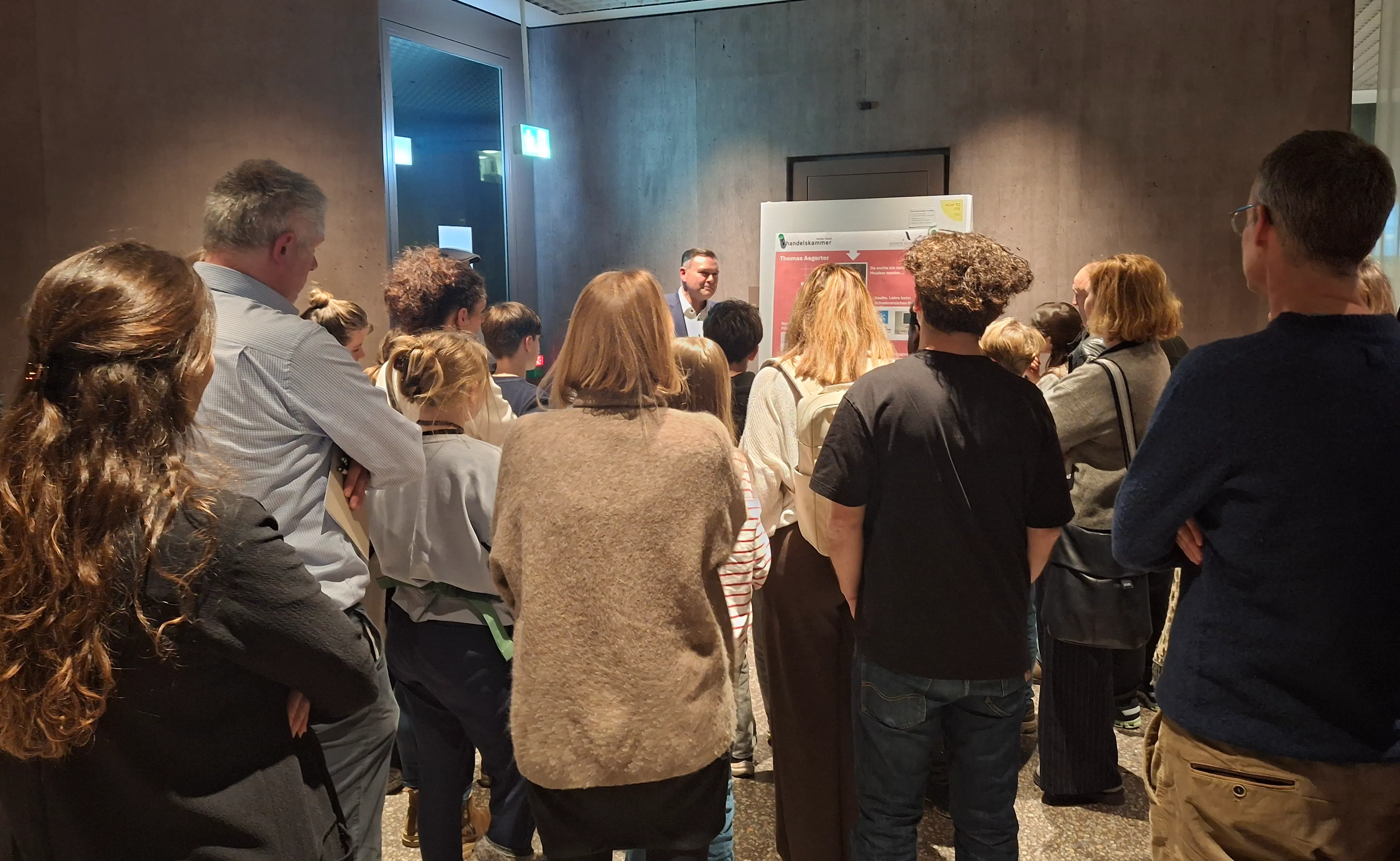 Das Bild zeigt Eltern und Kinder, die einem Mann vor seinem Plakatvortrag zuhören