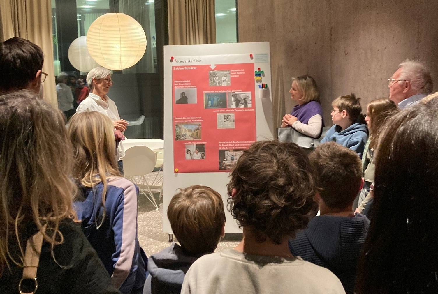 Das Bild zeigt Eltern und Kinder, die einem Mann vor seinem Plakatvortrag zuhören